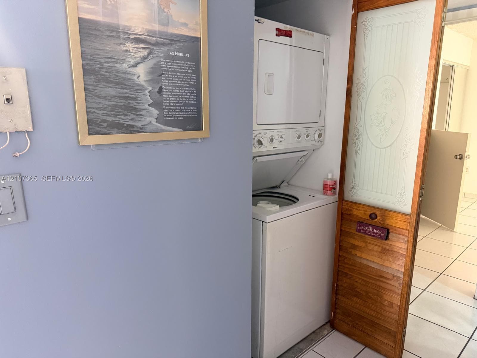 Stacked laundry unit featuring a white finish, enclosed by a wood-framed door with a frosted glass panel and decorative etching, set against tile flooring