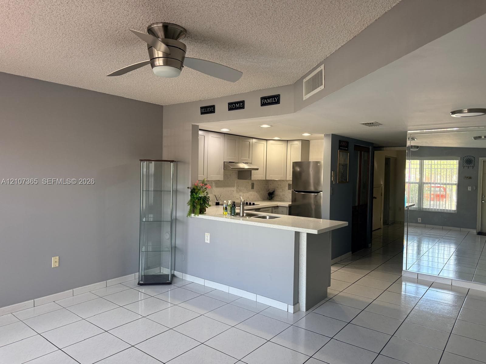 Open-concept space featuring white tile flooring, breakfast bar with stone-look countertop, stainless steel appliances, shaker-style cabinetry, and brushed nickel ceiling fan