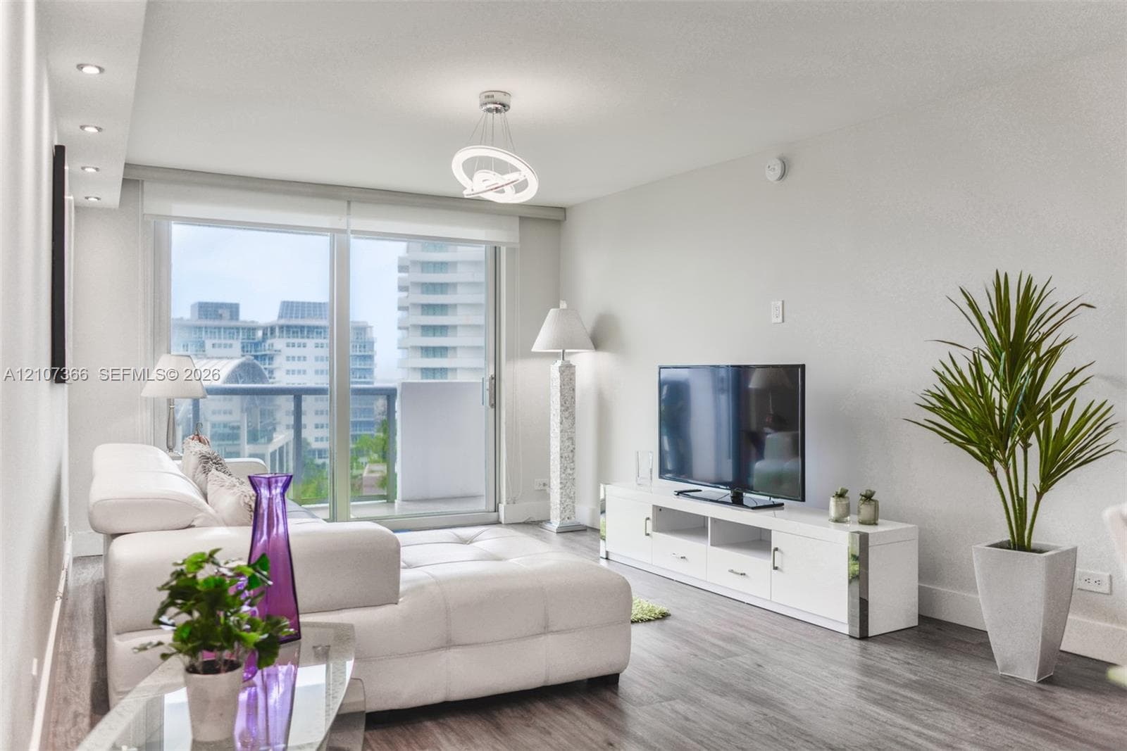 Open-concept living space featuring wood-finish flooring, a multi-tiered crystal chandelier, recessed lighting, and a kitchen with glass-front cabinetry and stone-tiled backsplash