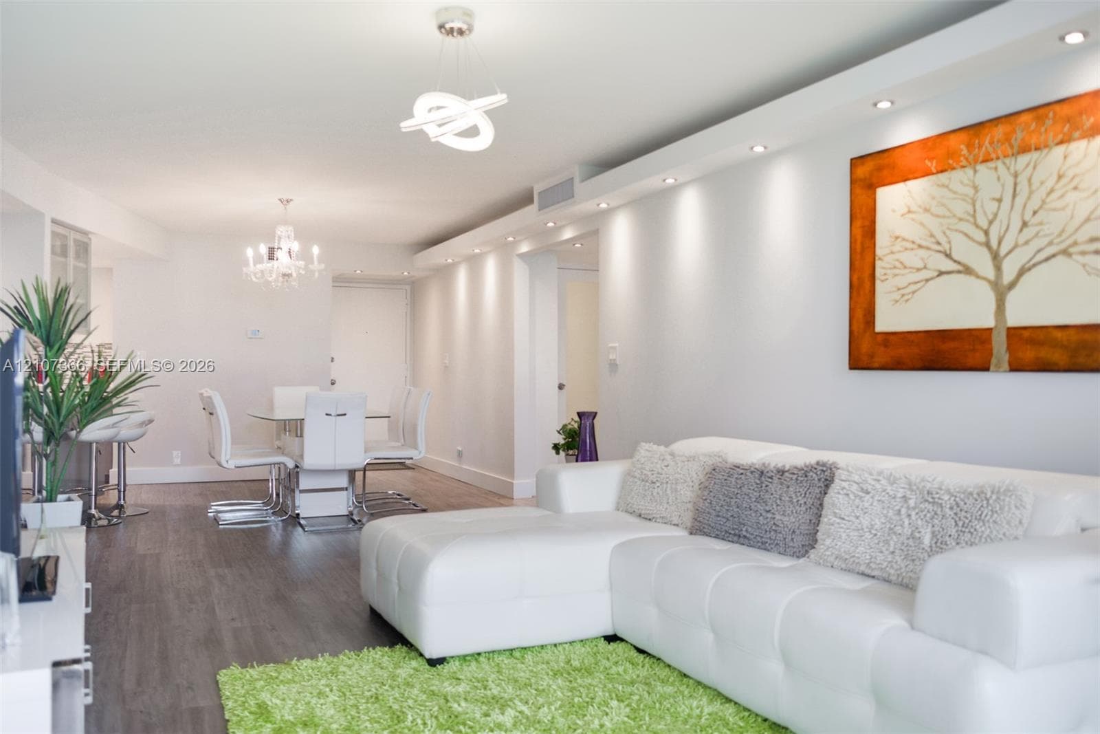 Open-concept interior featuring wood-finish flooring, recessed ceiling lighting, a structural soffit with integrated downlights, and contemporary geometric chandelier fixtures