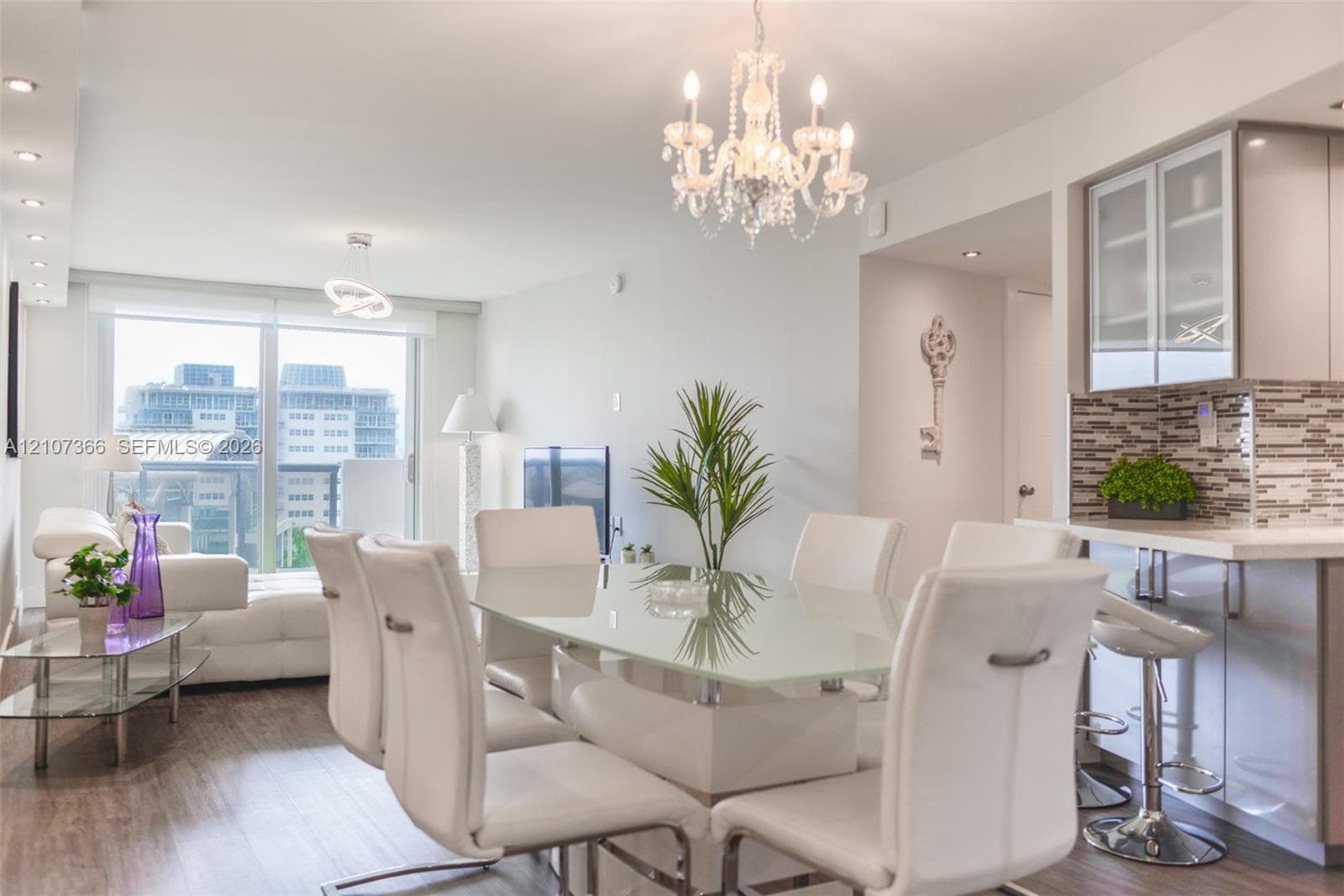 Open-concept living space featuring wood-finish flooring, a multi-tiered crystal chandelier, recessed lighting, and a kitchen with glass-front cabinetry and stone-tiled backsplash