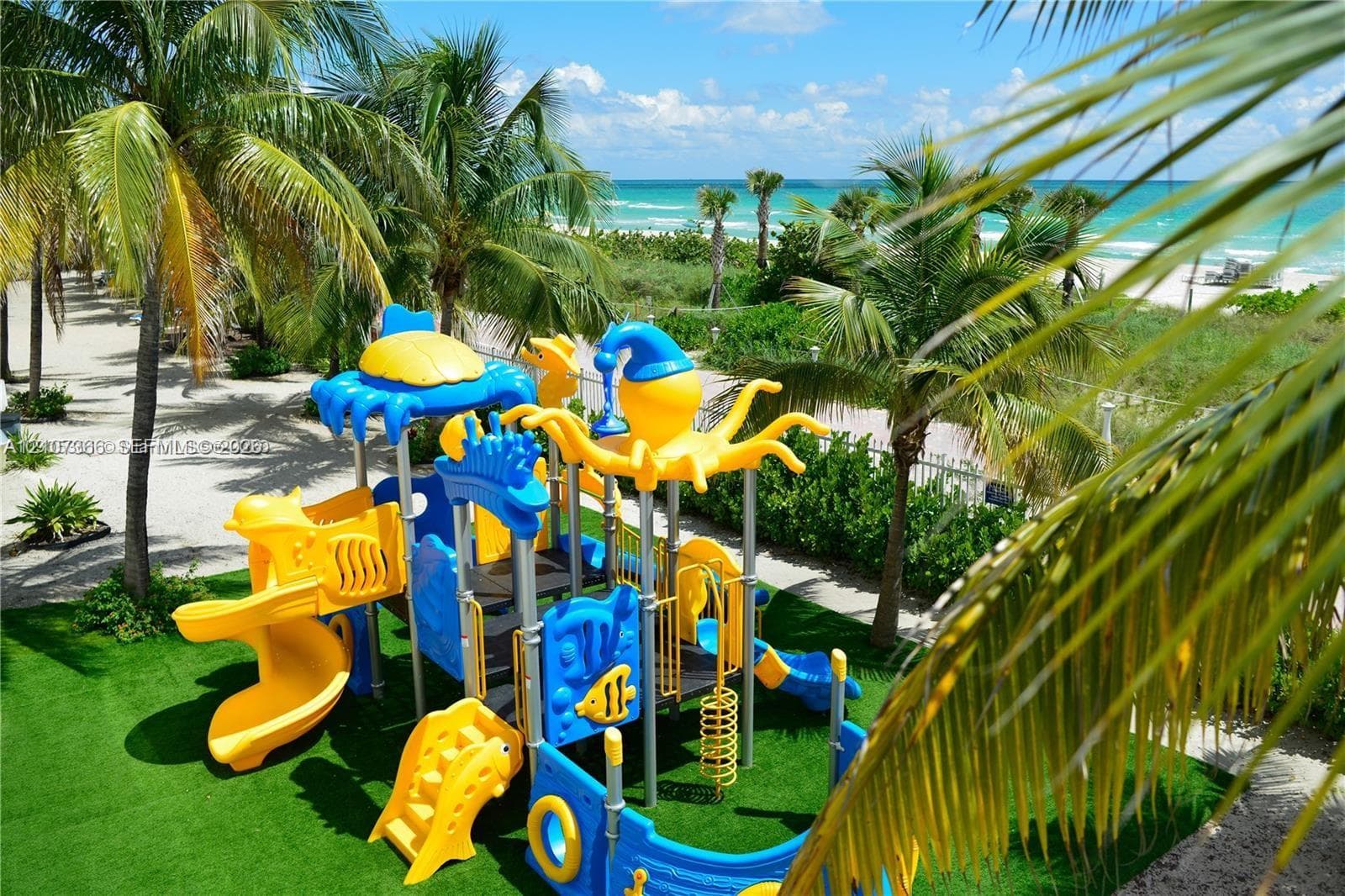 Outdoor play structure situated on artificial turf featuring mature palm trees and direct ocean proximity