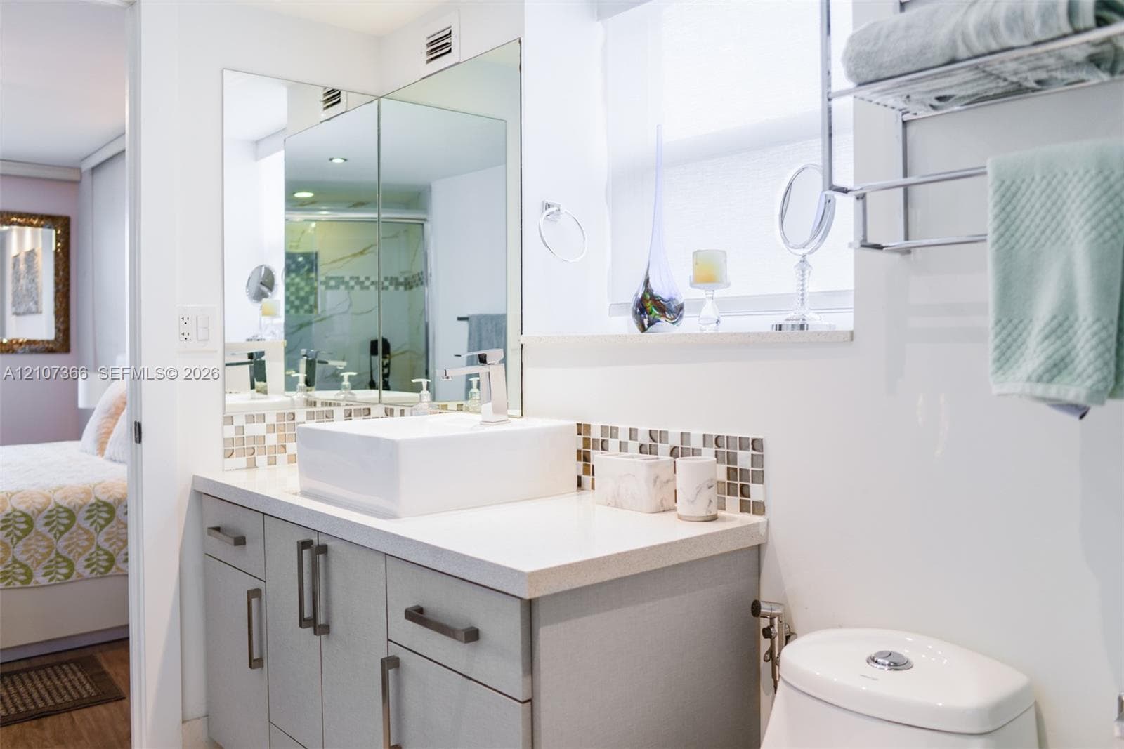 Bathroom vanity featuring a rectangular vessel sink, polished stone-surface countertop, mosaic tile backsplash, and light gray cabinetry with metallic hardware