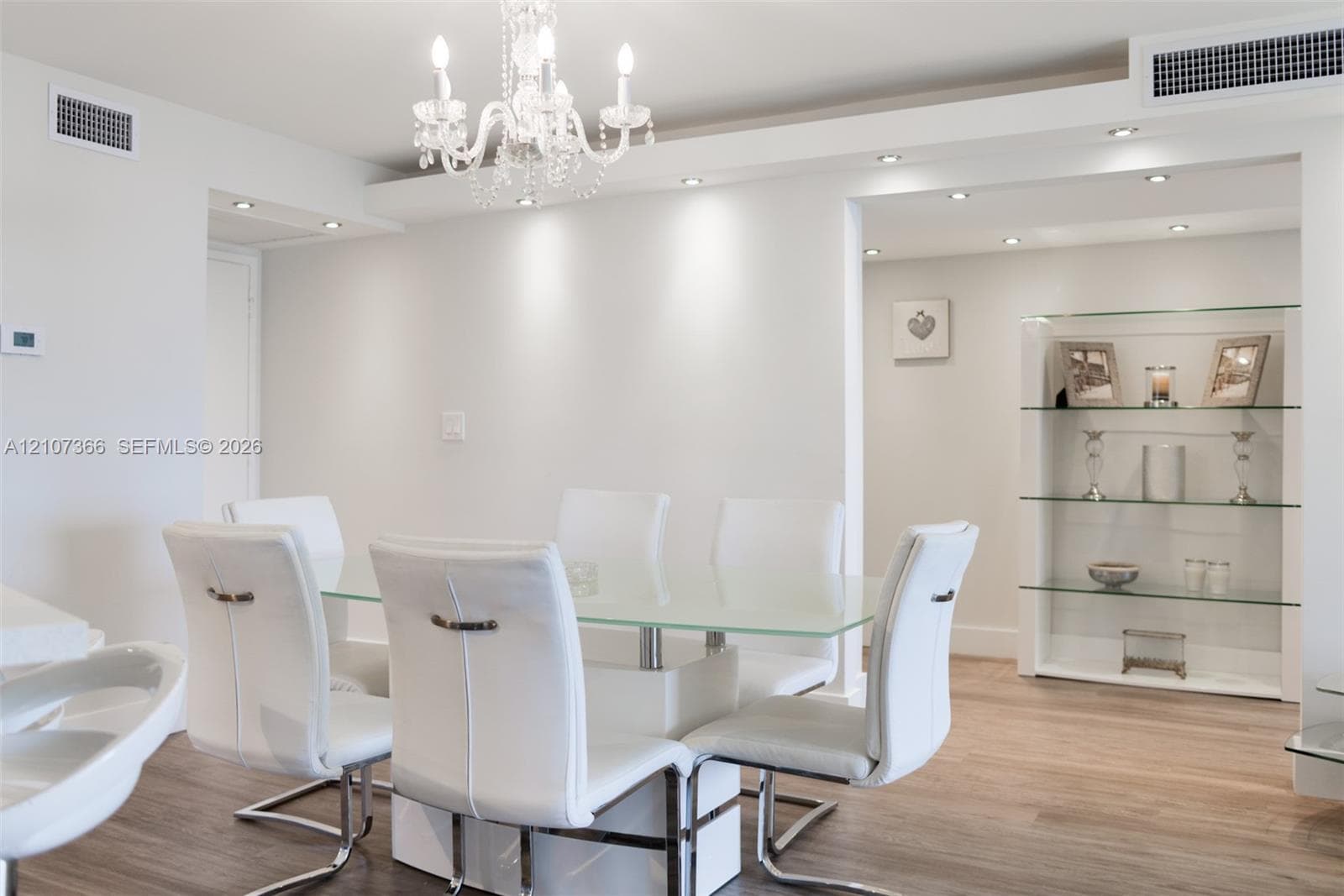Open-concept interior featuring wood-finish flooring, a multi-tiered crystal chandelier, recessed ceiling lighting, and a built-in floor-to-ceiling glass display shelving unit