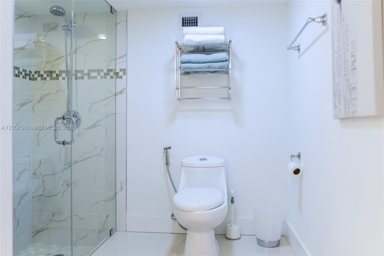 Walk-in glass-enclosed shower featuring large-format wall tiling with decorative mosaic accent, wall-mounted metal shelving unit, and bidet sprayer