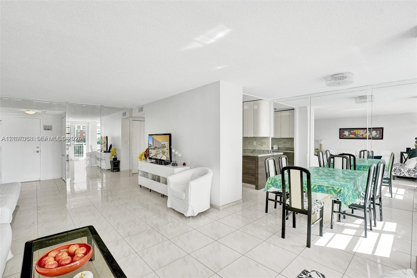 Open-concept floor plan featuring light-toned tile flooring, floor-to-ceiling mirror paneling, and a kitchen equipped with cabinetry and stone-surface counters