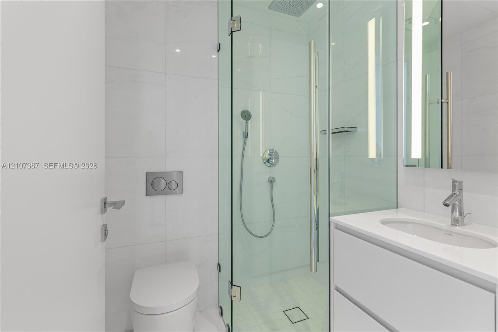 Bathroom featuring a glass-enclosed shower with a rainfall showerhead, wall-mounted dual-flush toilet, white vanity with an integrated sink, polished chrome fixtures, and large-format tile surround
