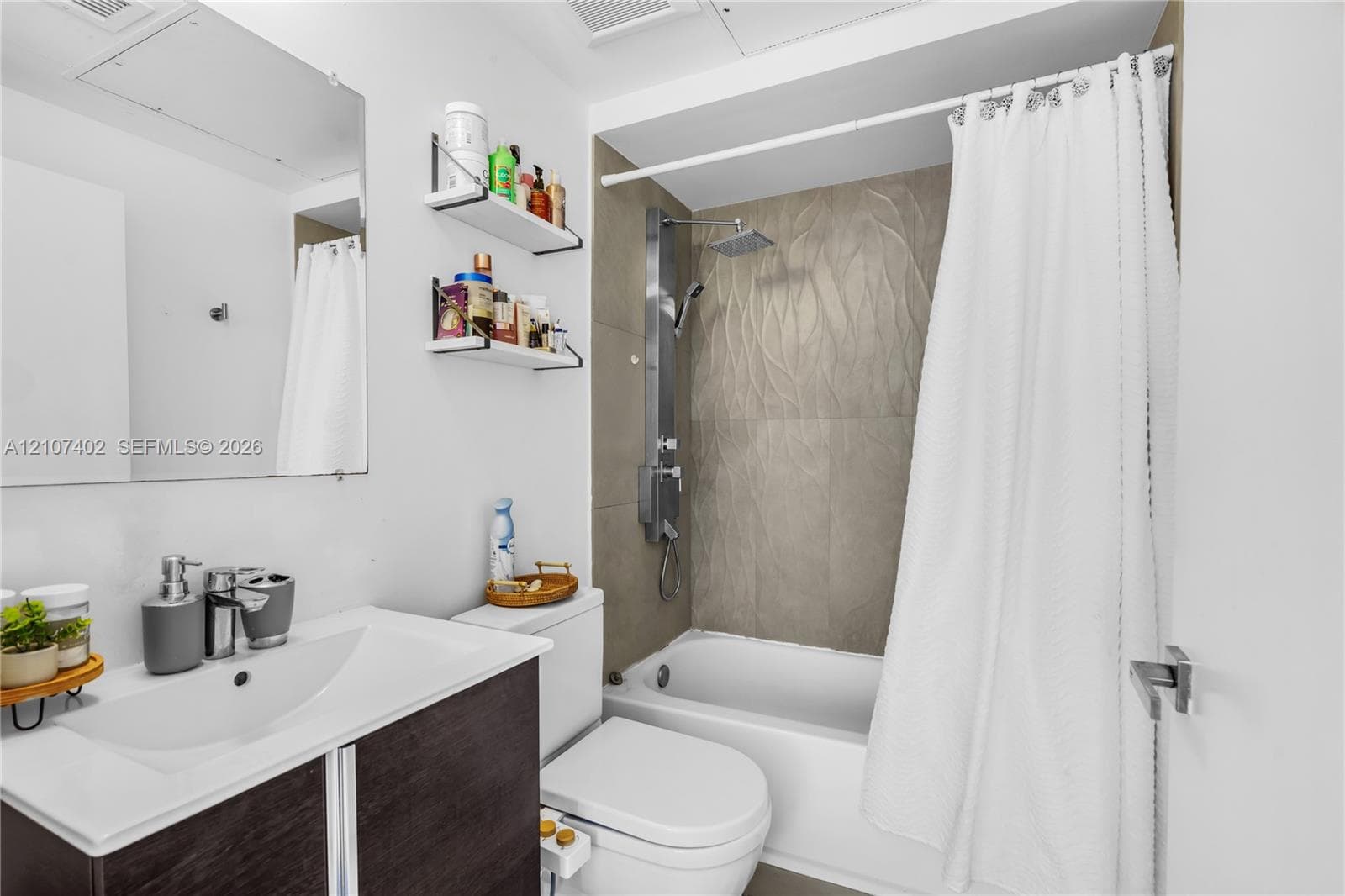 Bathroom featuring a dark wood-finish vanity with a white integrated basin, frameless wall-mounted mirror, two built-in wall shelves, a bathtub with textured tile surround, and a stainless steel shower panel system