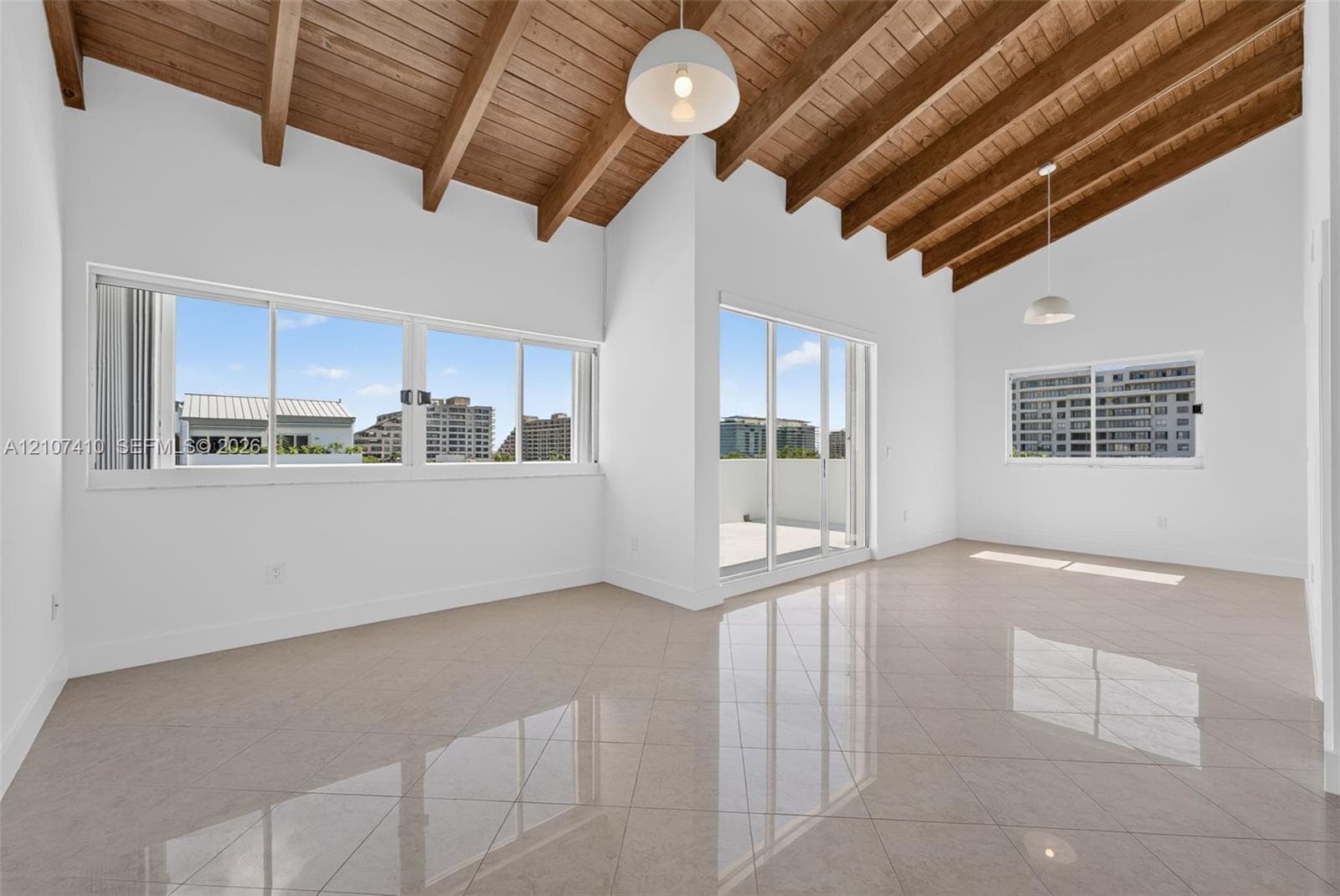 Vaulted ceilings with exposed wooden beams, large-format tile flooring, oversized windows, sliding glass door to a balcony, and dual pendant light fixtures