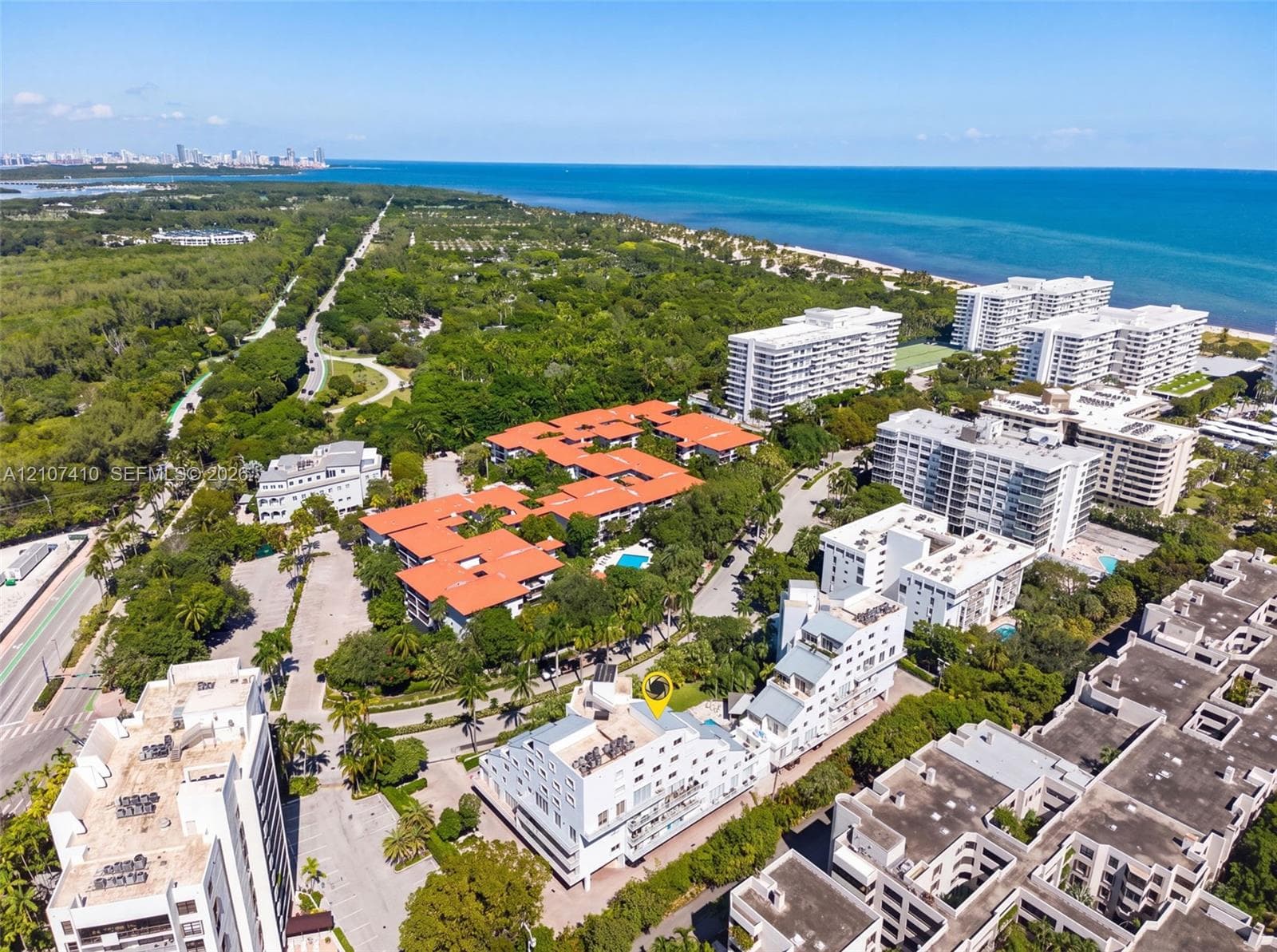 Aerial perspective of a multi-story residential complex featuring white stucco exterior finishes, flat roofing, and private balconies, situated in a coastal setting with proximity to wooded parkland and the shoreline