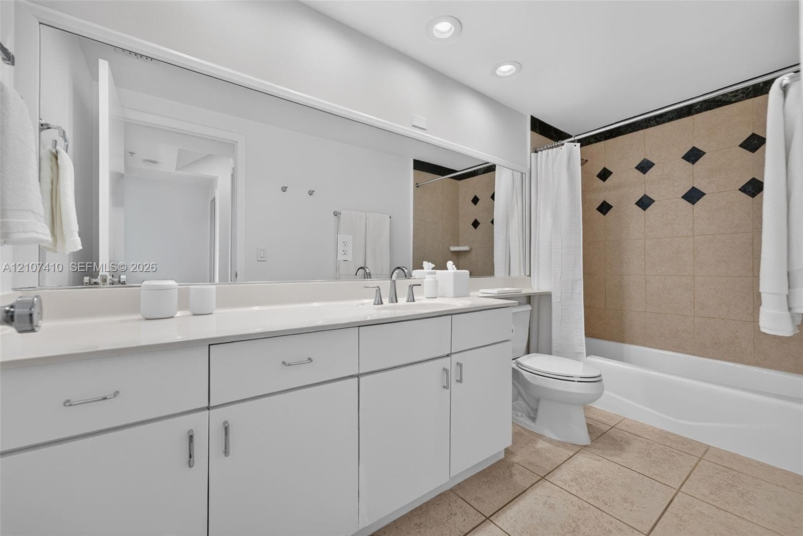 Bathroom featuring a vanity with white cabinetry and a solid-surface countertop, full-width wall mirror, recessed lighting, and a bathtub with tile surround and decorative insets