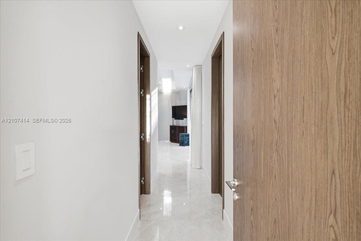 Hallway featuring large-format light-toned stone flooring, recessed ceiling lighting, and solid wood-finish doors with brushed-metal hardware