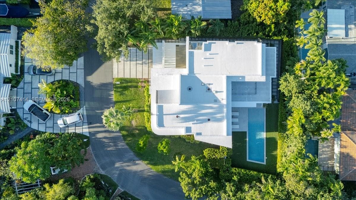 Aerial perspective of a multi-level residence featuring a rectangular swimming pool, a circular stone-paver driveway, mature tropical landscaping, and a flat roofline