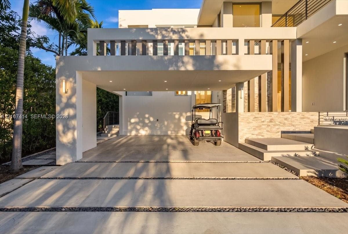 Modern architectural facade featuring a covered porte-cochère, recessed lighting, stacked stone exterior detailing, and a concrete driveway with decorative rock inlay