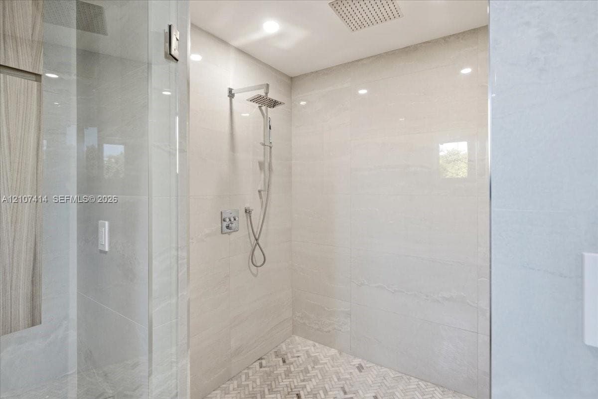 Walk-in shower featuring large-format stone wall tile, herringbone-patterned floor tile, a rainfall showerhead, a wall-mounted hand shower, and a glass enclosure