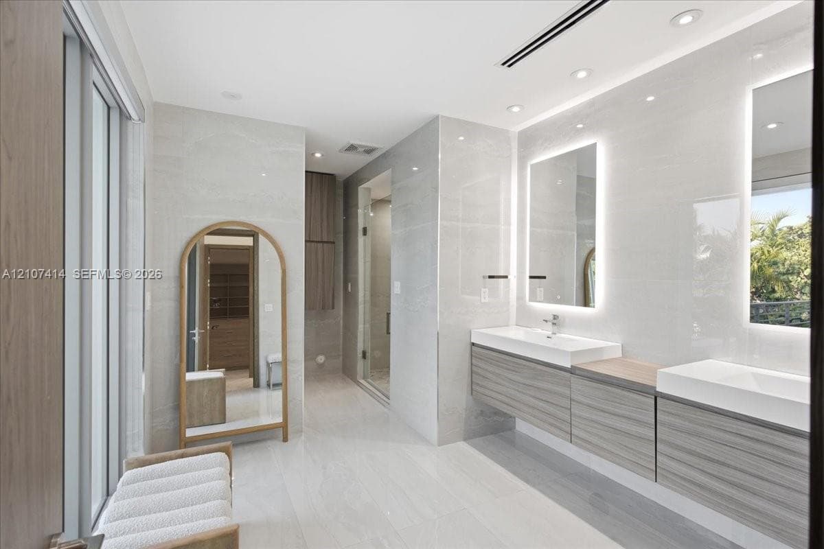Dual floating vanity with horizontal wood-finish cabinetry, integrated rectangular sinks, and backlit mirrors, accented by large-format neutral wall tiles and recessed lighting