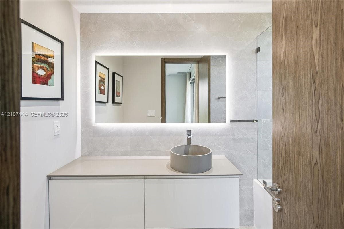 White vanity with a solid-surface countertop, cylindrical vessel sink, LED-backlit mirror, and full-height stone-finish wall tiling