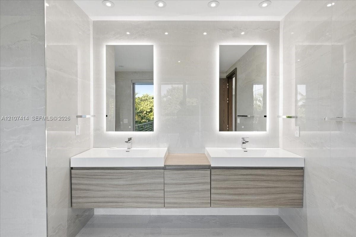Floating dual vanity with wood-finish cabinetry, white integrated sinks, backlit rectangular mirrors, and recessed ceiling lighting, set against large-format tiled walls