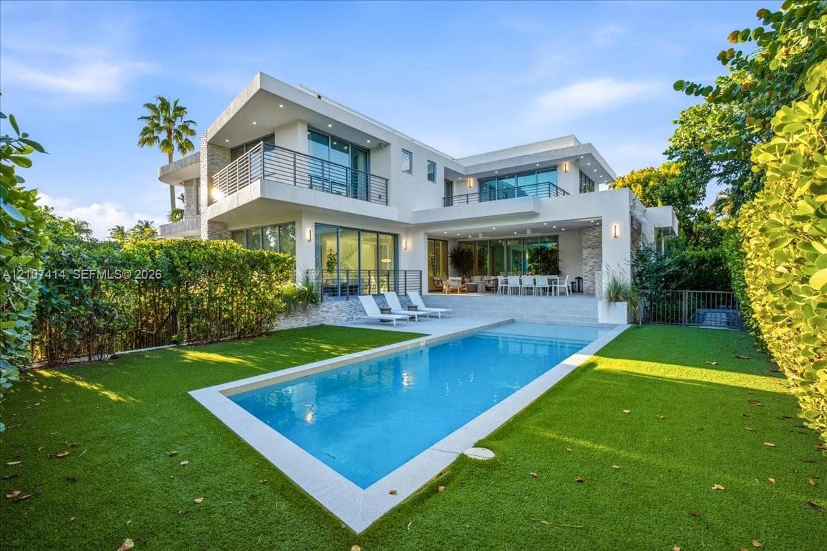 Modern two-story exterior featuring smooth stucco finish, stone accent pillars, expansive glass sliding doors, elevated balconies with metal railings, and a rectangular swimming pool surrounded by a manicured lawn