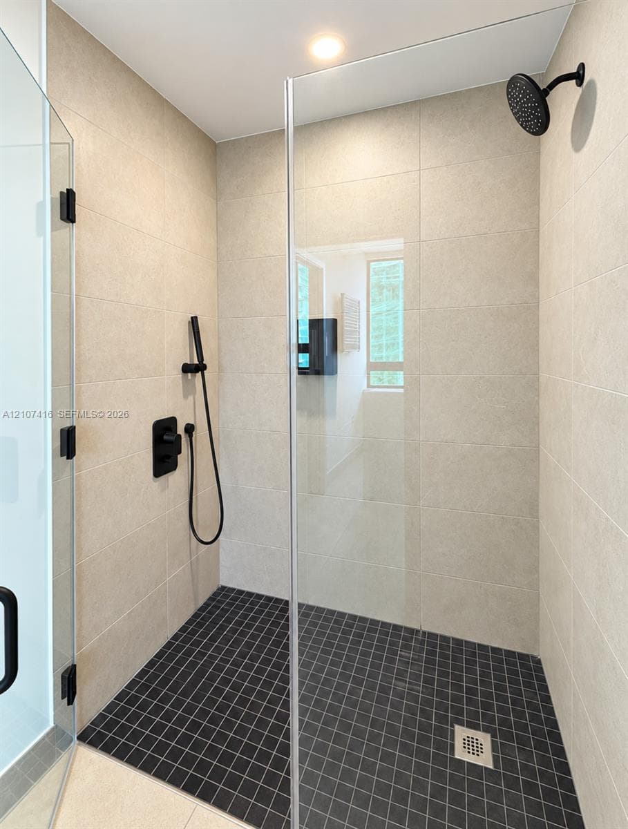 Walk-in shower featuring large-format wall tile, dark grid-patterned floor tile, frameless glass enclosure, and matte black plumbing fixtures including a rainfall showerhead and handheld wand