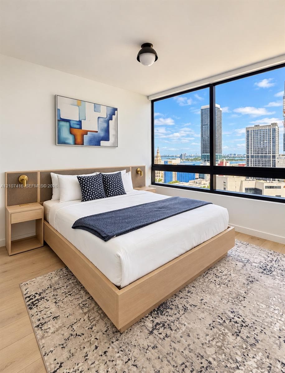 Floor-to-ceiling windows with expansive urban and water scenery, light wood-finish flooring, built-in wooden headboard with integrated side tables, and matte brass wall-mounted light fixtures