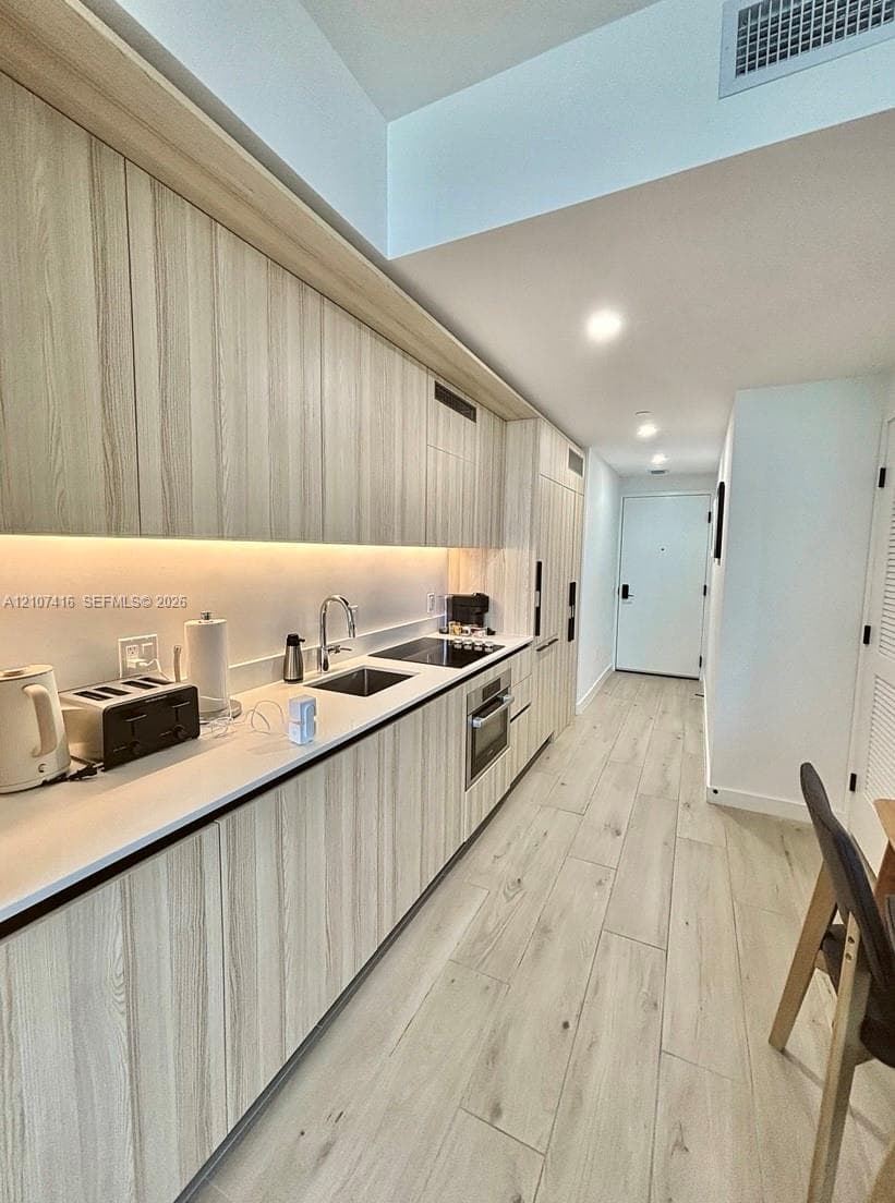 Kitchen featuring light wood-finish cabinetry, solid surface countertops, under-cabinet lighting, integrated appliances, and wood-finish flooring