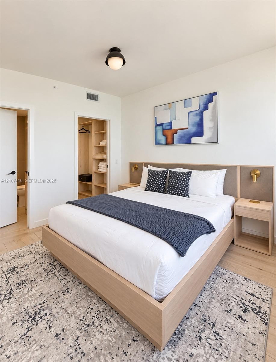 Light-toned wood-finish flooring, built-in light-toned wood headboard with integrated nightstands, brushed brass wall-mounted light fixtures, and walk-in closet with custom shelving