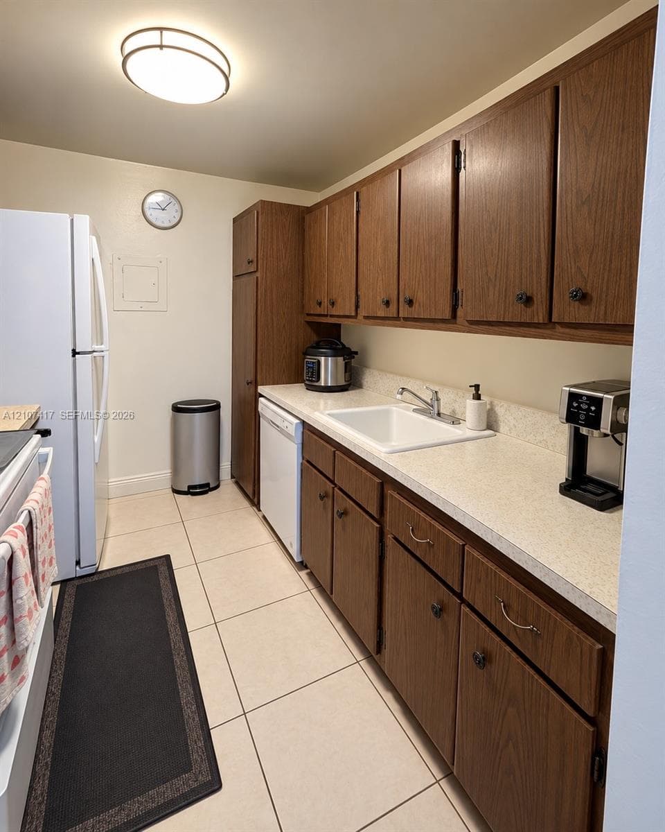 Kitchen featuring wood-finish cabinetry, neutral solid-surface countertops, a porcelain sink with a chrome faucet, tile flooring, and a flush-mount overhead light fixture