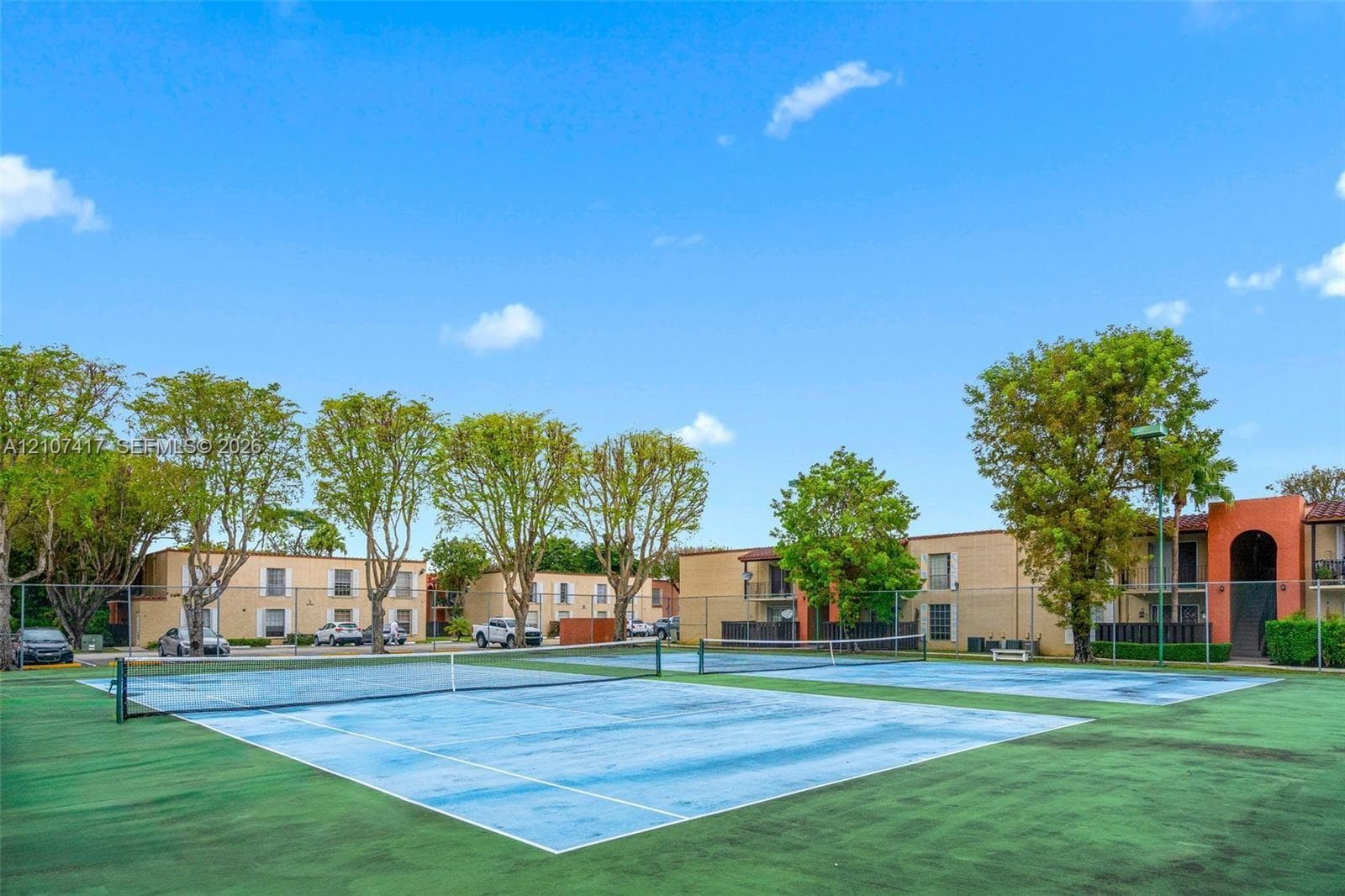 Dual tennis courts featuring hard-surface playing areas, perimeter chain-link fencing, and mature surrounding landscaping