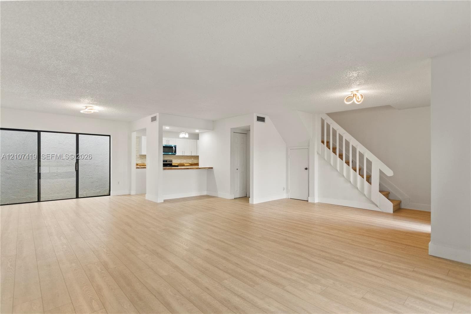 Open-concept interior featuring light wood-finish flooring, sliding glass door, white cabinetry, built-in breakfast bar, and a staircase with white balusters