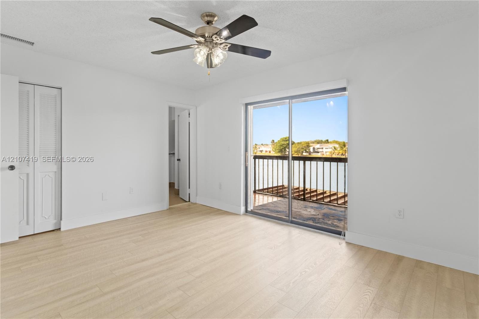 Interior space featuring light wood-finish flooring, a ceiling fan with integrated lighting, louvered-panel bi-fold doors, and a sliding glass door providing access to a railed balcony