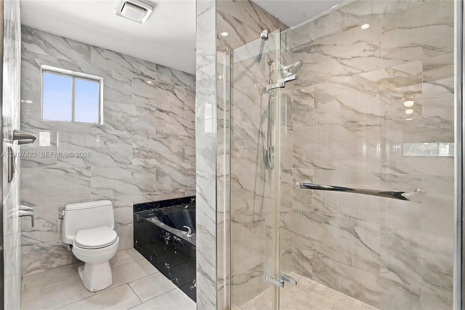 Bathroom featuring floor-to-ceiling veined tile, a glass-enclosed shower, a black stone-finish bathtub surround, and a white porcelain toilet