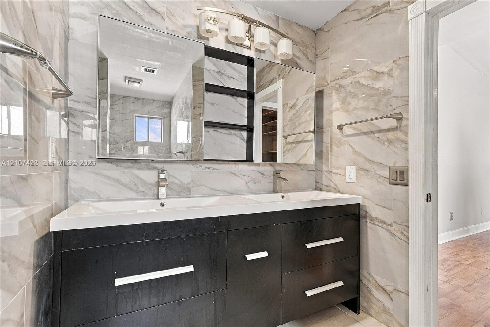 Double vanity featuring a dark-toned cabinet base, integrated dual sinks, large-scale tile wall surfacing, polished metal hardware, and multi-light vanity fixtures