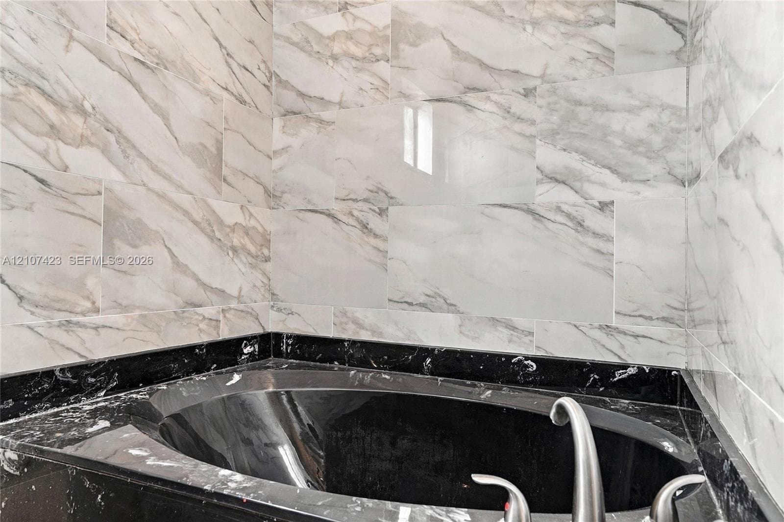 Built-in soaking tub with a dark stone-finish surround, complemented by floor-to-ceiling wall tile featuring a veined pattern and a metallic faucet fixture