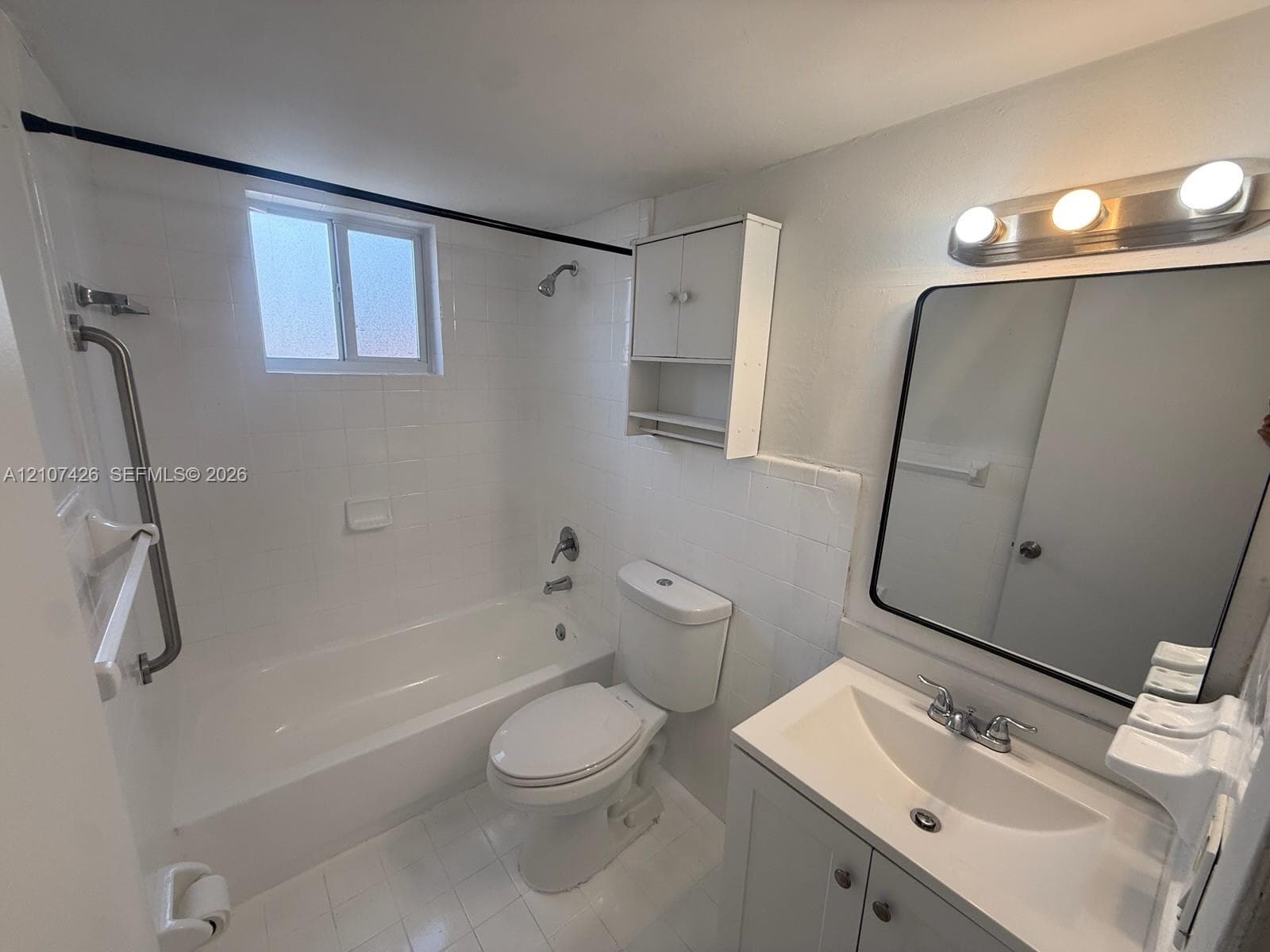 Bathroom featuring a white vanity with an integrated sink, a rectangular mirror with a black frame, a three-light vanity fixture, white tile wall surfacing, and a built-in wall cabinet