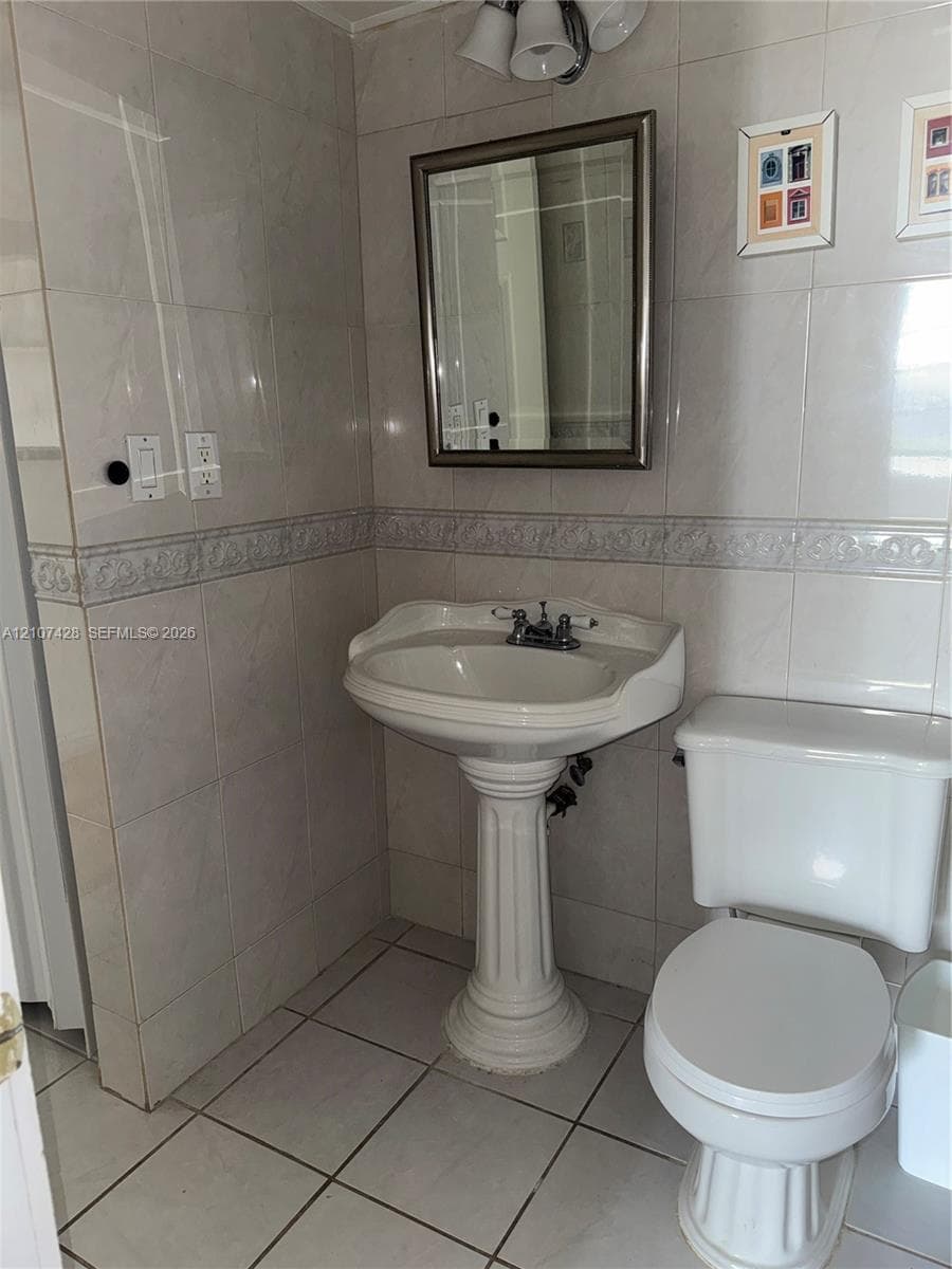 Full-wall tile surround with decorative accent border, white pedestal sink, polished chrome plumbing fixtures, rectangular wall-mounted mirror, and tile flooring