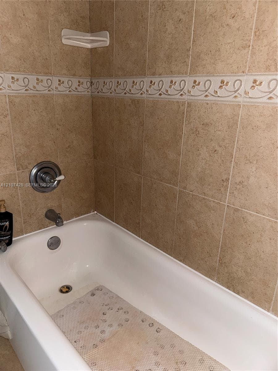 Bathtub featuring wall-mounted chrome fixtures and full-height stone-finish tile surround with an integrated decorative border and built-in corner shelf
