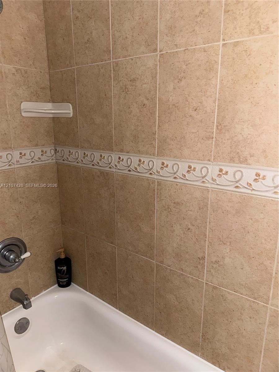 Bathtub surround featuring neutral-toned square wall tiles, a decorative floral border inlay, integrated corner soap dish, and chrome-finish plumbing fixtures
