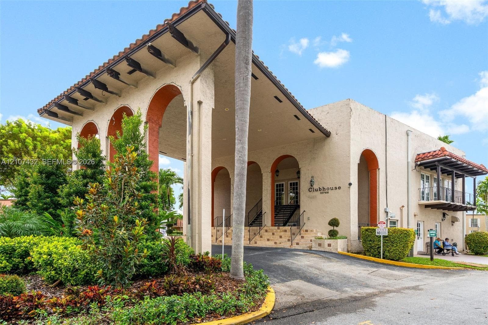Clubhouse exterior featuring a covered porte-cochère, decorative archways, stucco facade, and a second-story balcony with a tiled roof overhang