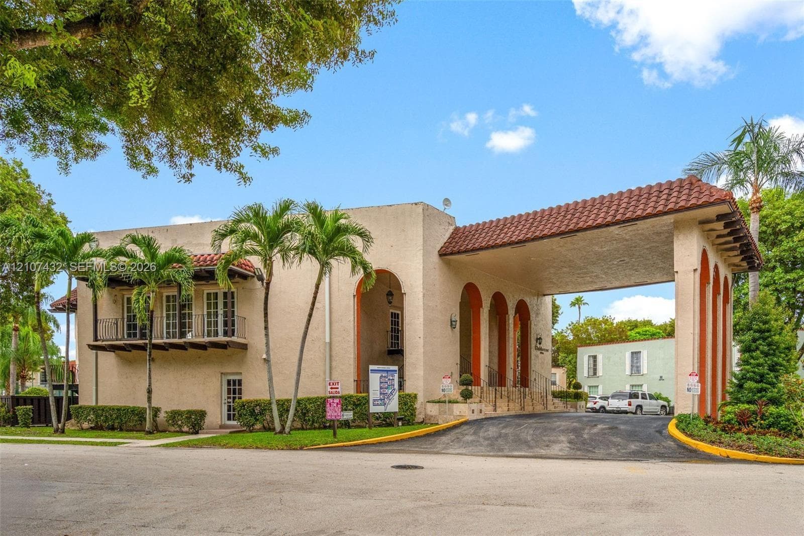 Stucco exterior featuring a large covered porte-cochère, arched architectural detailing, a second-story balcony with metal railing, and mature palm landscaping