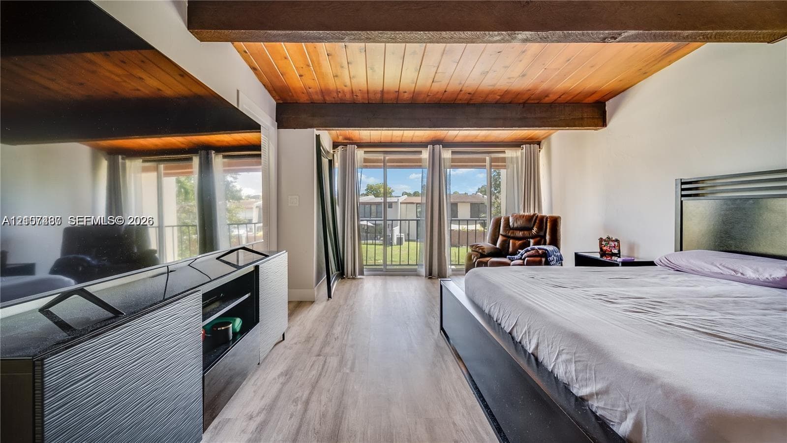 Wood-plank ceiling with exposed timber beams, light-toned wood-finish flooring, and floor-to-ceiling glass sliding doors opening to an exterior balcony