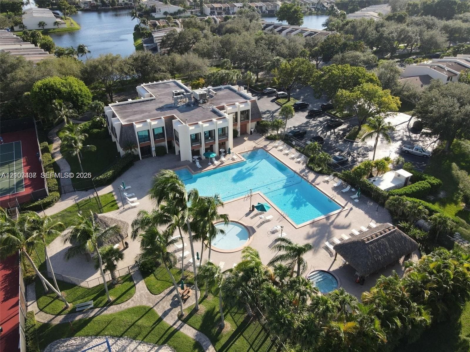 Aerial perspective of a multi-level complex featuring a swimming pool, separate spa, tennis court, and a thatched-roof cabana structure, situated within landscaped grounds