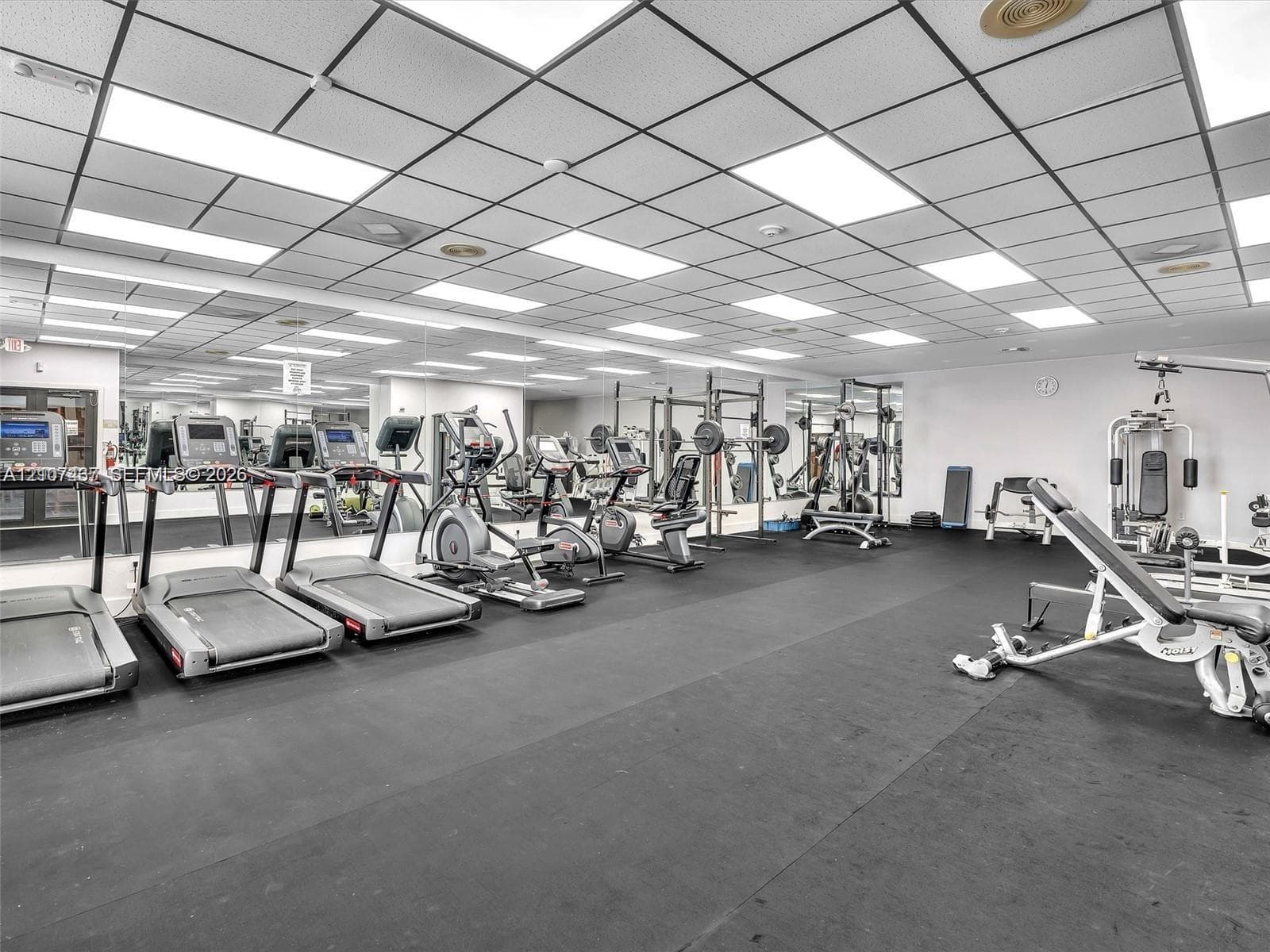 Fitness room featuring rubberized flooring, a drop ceiling with integrated fluorescent lighting, and wall-to-wall mirrors