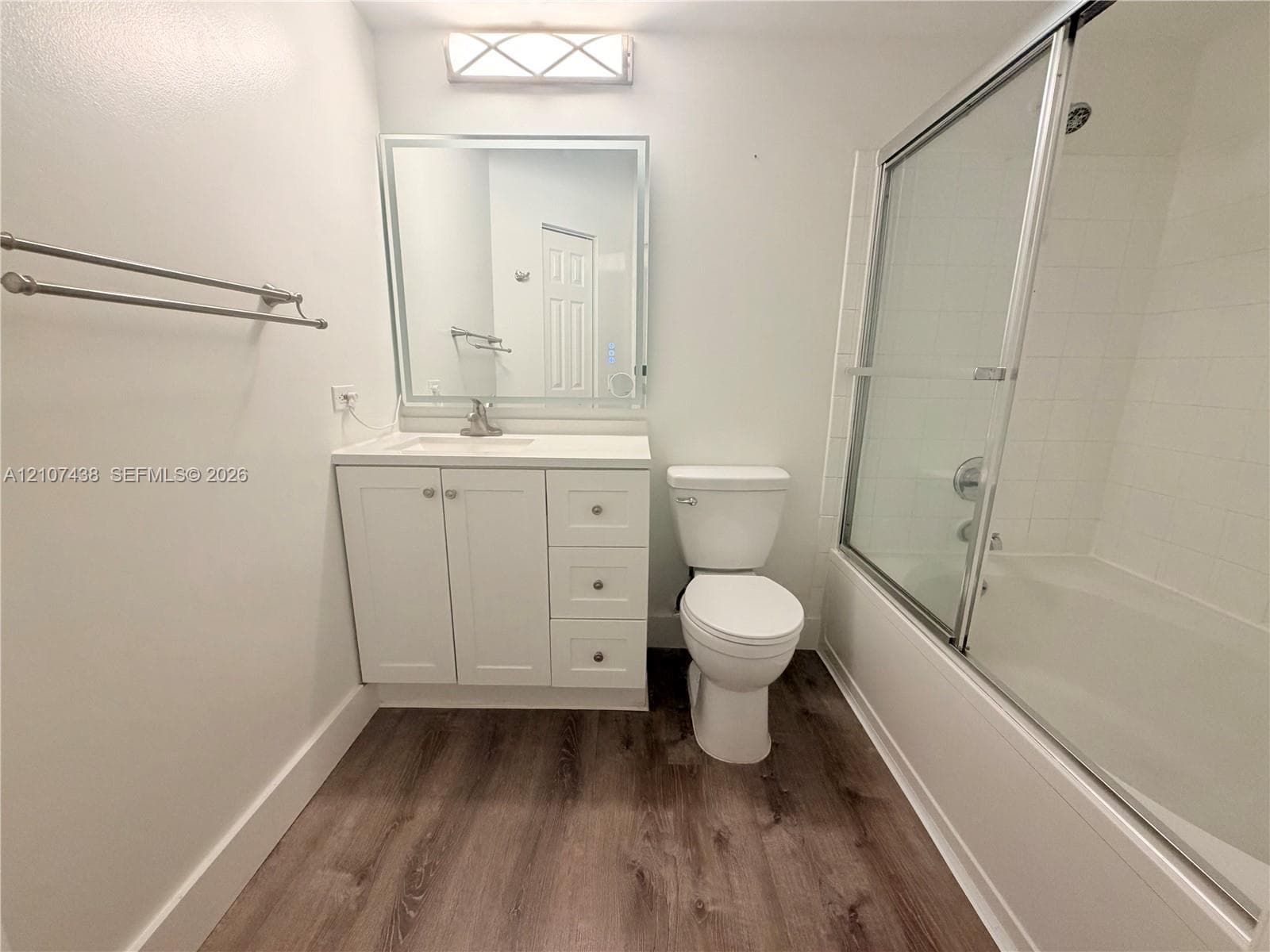 White vanity with a rectangular sink, illuminated mirror, wood-finish flooring, and a shower-tub combination with glass sliding doors