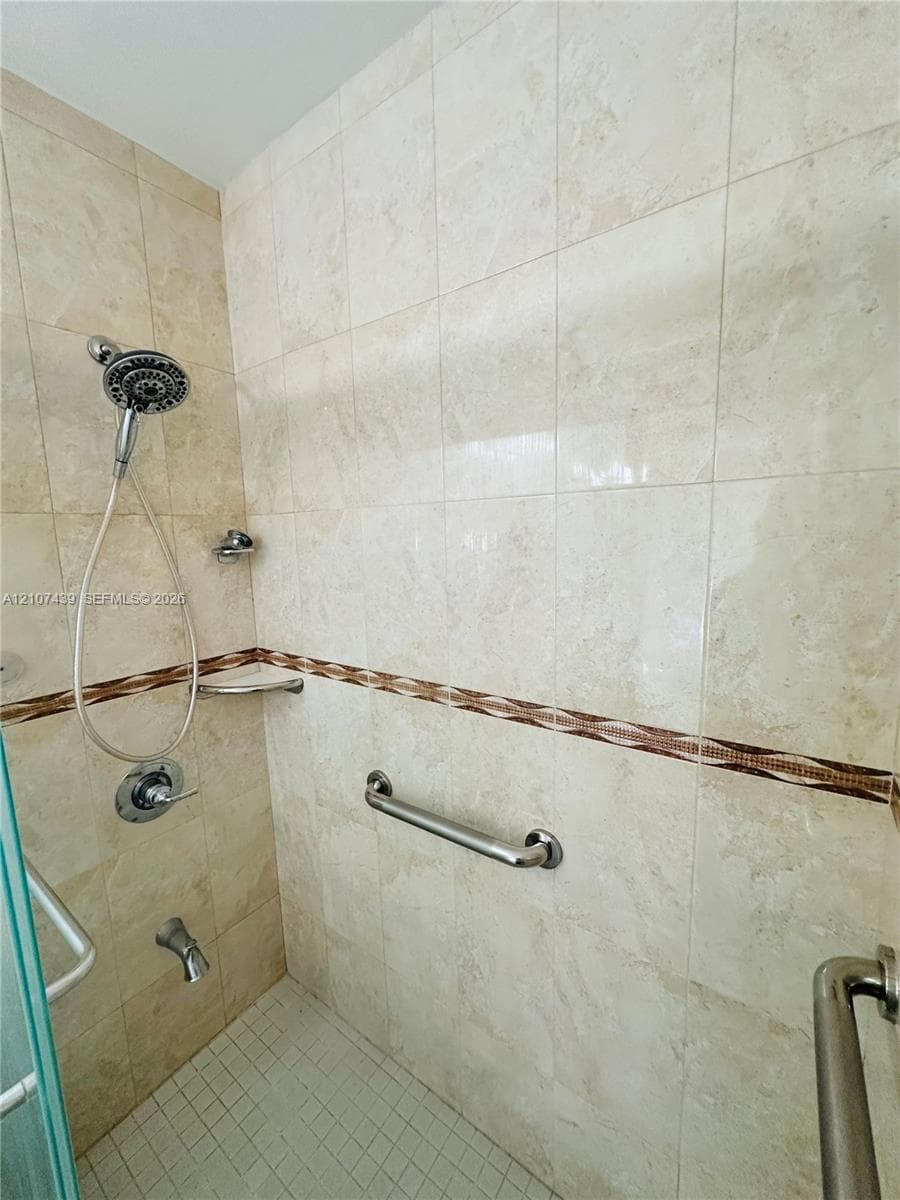 Walk-in shower featuring floor-to-ceiling neutral tile, a decorative mosaic border, a multi-setting handheld showerhead, integrated corner shelving, and metal support rails