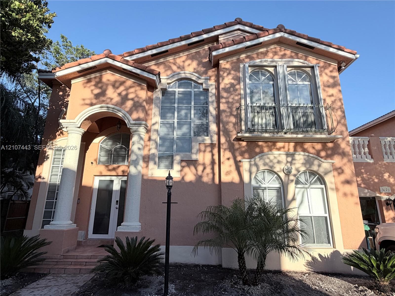 Stucco exterior featuring dual classic columns, arched multi-pane windows, a decorative metal balcony, and a multi-tiered roofline