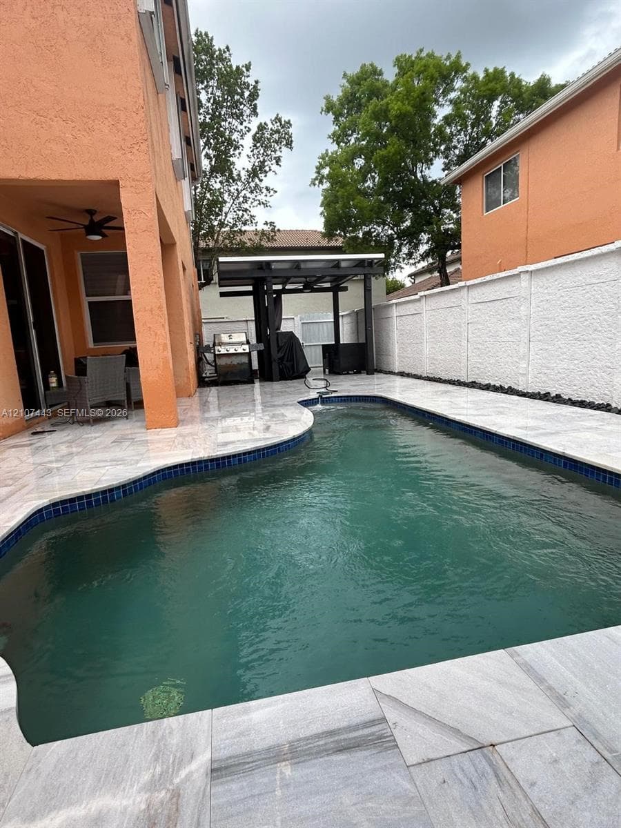 Outdoor swimming pool featuring stone-finish decking, covered patio space with ceiling fan, and privacy wall