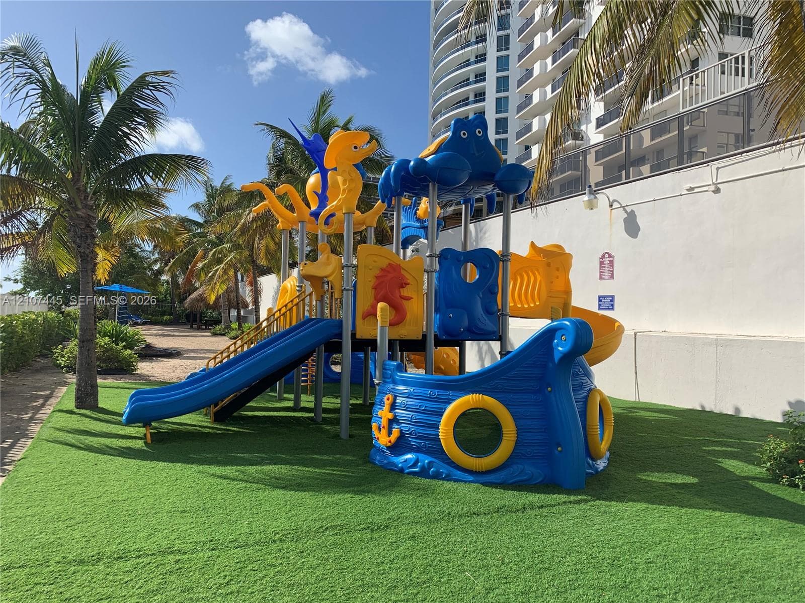Outdoor recreational area featuring a multi-level playground structure, synthetic turf ground cover, and mature palm trees