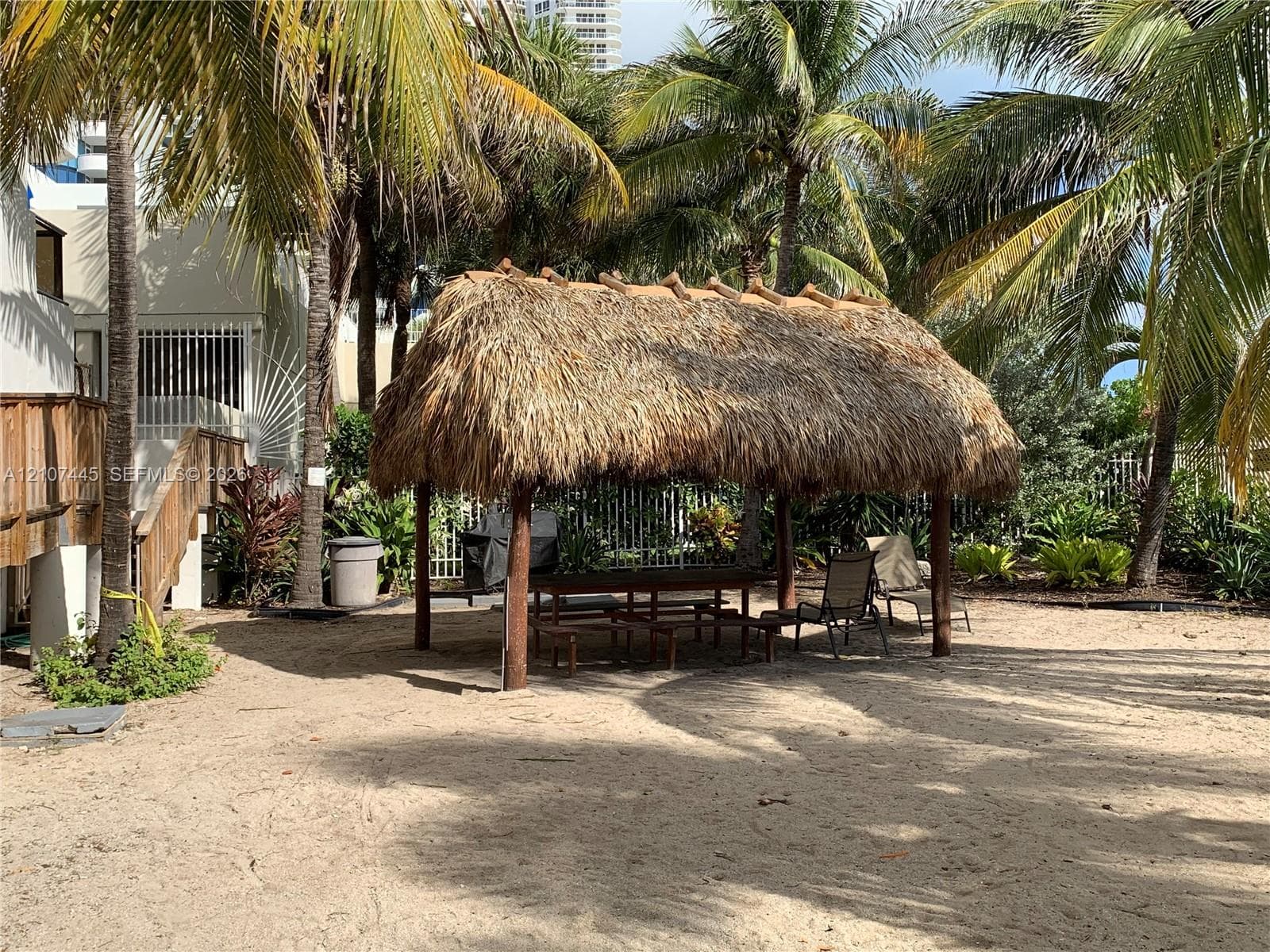 Outdoor area featuring a thatched-roof tiki hut, sandy ground surface, and mature palm trees