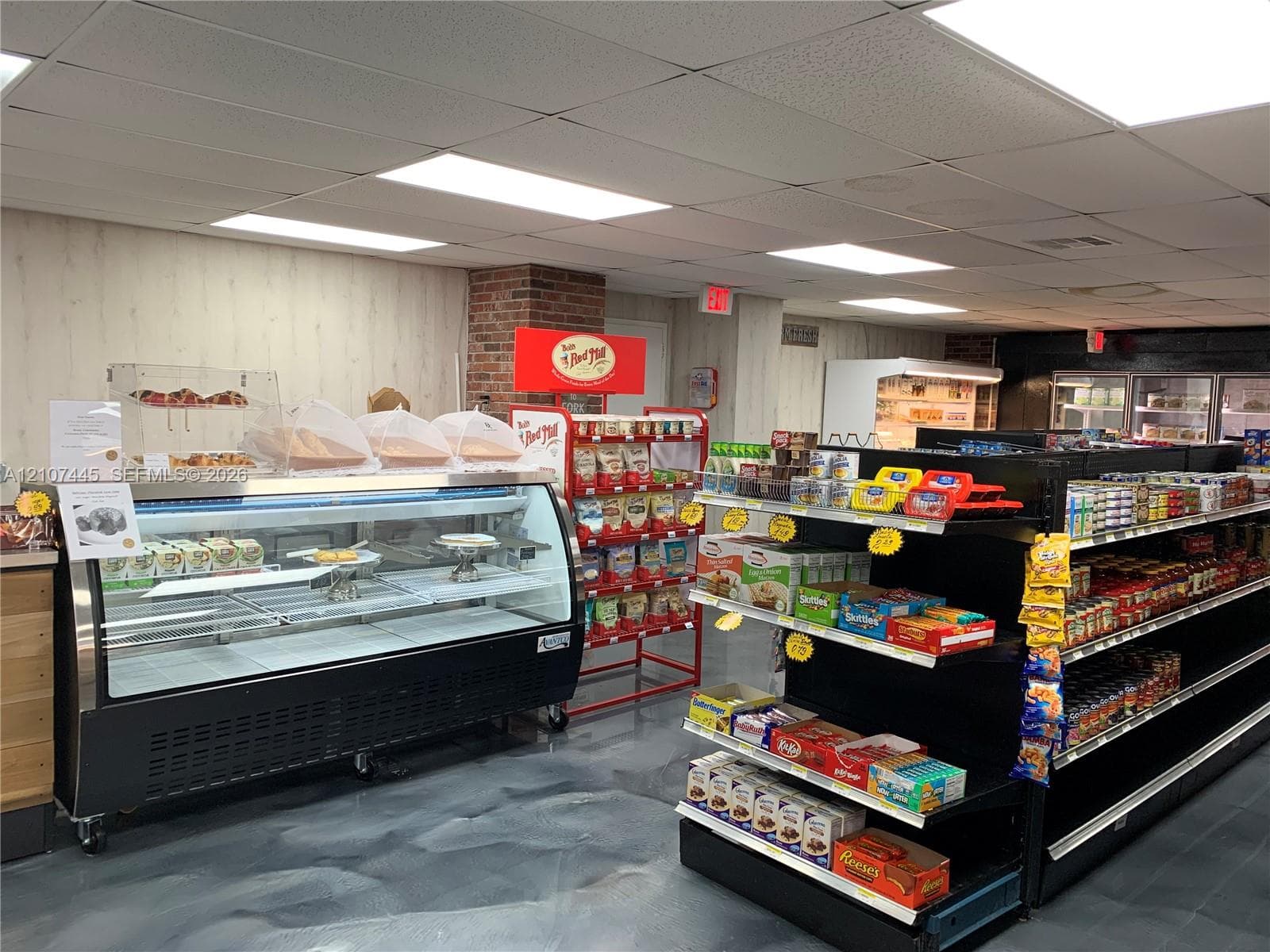 Commercial interior featuring a refrigerated display case, polished concrete flooring, recessed drop ceiling lighting, exposed brick accent pillar, and wood-paneled wall finish
